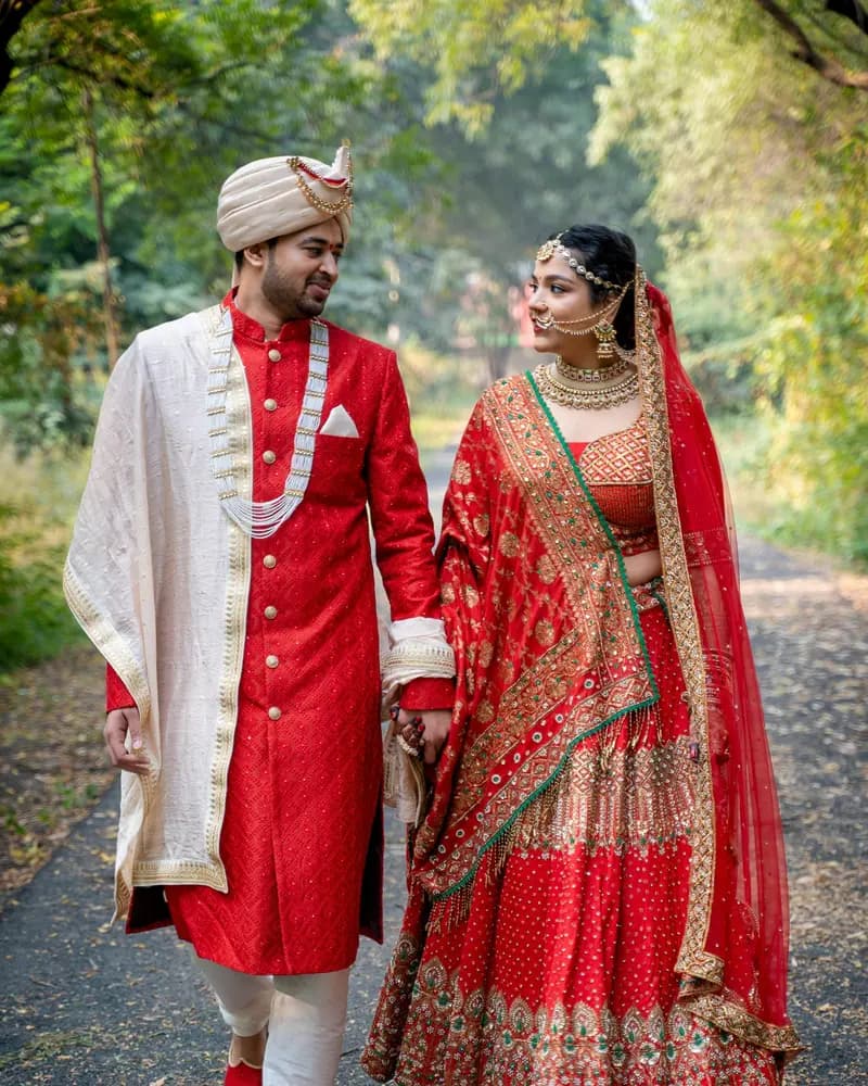 A bride and groom in red and ivory walking hand in hand along a tree-lined path